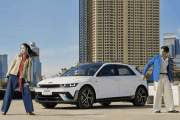 White compact car parked on a sunny rooftop with a man in a blue blazer and a woman in a tan coat posing beside it against a city skyline.