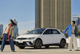 White compact car parked on a sunny rooftop with a man in a blue blazer and a woman in a tan coat posing beside it against a city skyline.