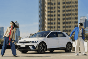 White compact car parked on a sunny rooftop with a man in a blue blazer and a woman in a tan coat posing beside it against a city skyline.