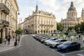 Cobbled city street with historic buildings and a row of electric cars parked at charging stations on the right side, near a café on the left.