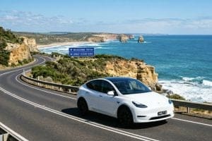 White electric car on a winding coastal road with rocky cliffs and the blue ocean; road signs indicate Melbourne 150 km and the Great Ocean Road.