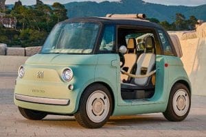 Mint green vintage Fiat 500 with the driver's door open, parked on a stone plaza with hills in the background.