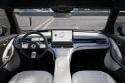 Interior of a modern car showing a large central touchscreen, steering wheel, and light-gray seats in view of the dashboard and windshield outside.