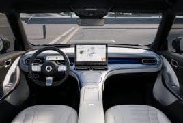 Interior of a modern car showing a large central touchscreen, steering wheel, and light-gray seats in view of the dashboard and windshield outside.
