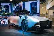 Electric sports coupe on display in a showroom with a large LED wall behind and a row of chairs nearby, spotlighting its sleek lines.
