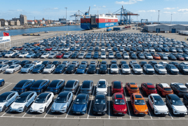 Rows of parked cars at the Port of Bremerhaven, with a cargo ship and cranes in the harbor behind them.