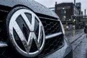 Front grille of a VW car with a large chrome VW emblem, raindrops on the surface and industrial buildings in the background.