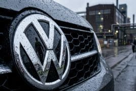 Front grille of a VW car with a large chrome VW emblem, raindrops on the surface and industrial buildings in the background.