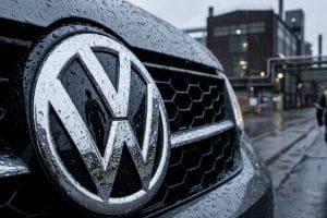 Front grille of a VW car with a large chrome VW emblem, raindrops on the surface and industrial buildings in the background.