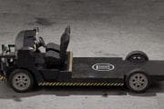 Industrial tow tractor with open cockpit and single seat on a flatbed platform, parked on a concrete floor with yellow-black safety stripes along the base.