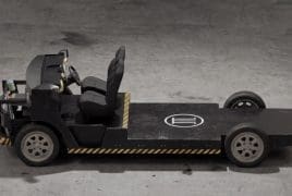 Industrial tow tractor with open cockpit and single seat on a flatbed platform, parked on a concrete floor with yellow-black safety stripes along the base.