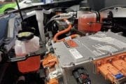 Under-hood view of an electric vehicle powertrain: orange high-voltage cables, a metal inverter housing, and coolant and brake fluid reservoirs visible.