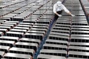 Worker in a white cleanroom suit, mask, and gloves inspecting rows of long battery modules arranged on racks in a manufacturing facility.