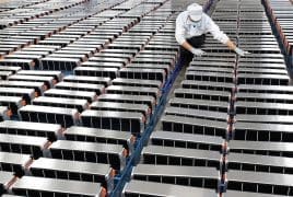 Worker in a white cleanroom suit, mask, and gloves inspecting rows of long battery modules arranged on racks in a manufacturing facility.