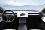 Front-seat view of a modern car interior with a large central touchscreen displaying a car image, a round steering wheel, and a mountain landscape visible through the windshield.