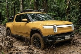 Yellow Rivian pickup driving through a muddy forest trail with a roof rack and muddy tires (off-road scene).