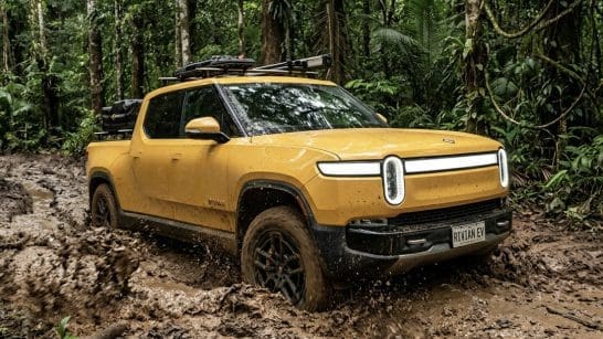 Yellow Rivian pickup driving through a muddy forest trail with a roof rack and muddy tires (off-road scene).