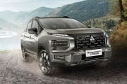 Silver Mitsubishi Xpander Cross SUV driving on a dirt road beside a lake with mountains in the background, overcast sky above the scene.