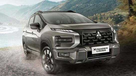 Silver Mitsubishi Xpander Cross SUV driving on a dirt road beside a lake with mountains in the background, overcast sky above the scene.